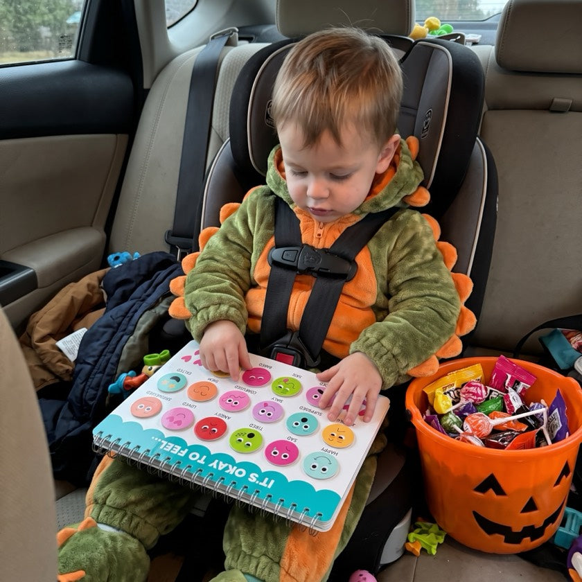A toddler in a dinosaur costume in a car seat with a Hadley Busy Book and a pumpkin candy bucket